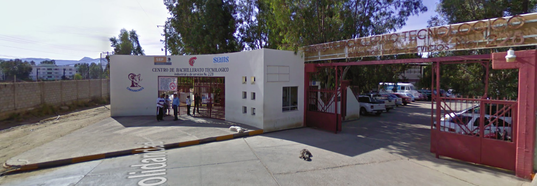 Fotografía en la que se muestra la entrada del plantel. Del lado izquierdo hay unos muros blancos y en medio de ellos está una reja color rojo por la que se puede ingresar al plantel. Del lado derecho hay un portón color rojo por el que pueden ingresar autos. 